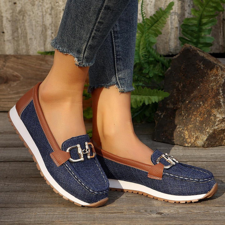 Navy slip-on loafers with brown strap, gold chain bar detail, and white rubber sole.